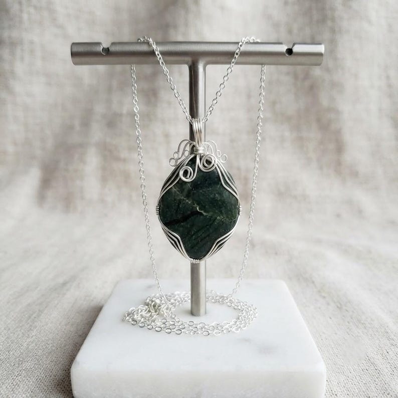Natural certified green jade pendant displayed as a handcrafted wire-wrapped necklace with a polished crystal body, custom silver-tone wire framing, and neat loop details. The dark green stone provides a focal point while the artistic wire wrapping adds a refined decorative character. This unique necklace is suitable for crystal lovers, gemstone collectors, spiritual jewelry styling, statement accessories, special occasions, and thoughtful holiday or birthday gifts with an organic feel.