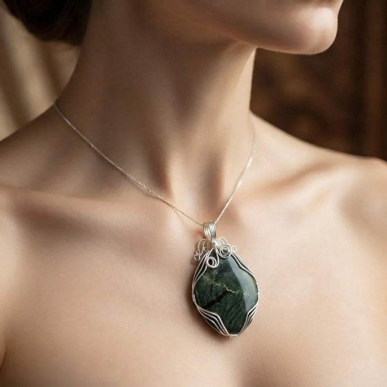 Handmade certified green jade crystal jewelry featuring a wire-wrapped pendant with a 90-100 carat stone base, smooth polished edges, and a decorative silver wire loop bail. The distinctive pendant design combines crystal beauty with refined wirework craftsmanship, creating an elegant statement necklace. Suitable for casual fashion, spiritual styling, elegant outfits, gemstone collections, crystal displays, and special gifts for women who appreciate fine and symbolic accessories.