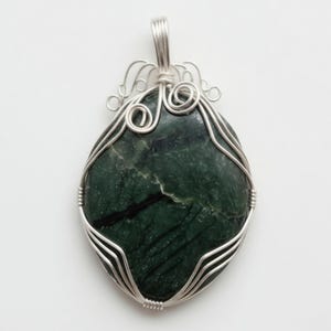 Unique certified green jade wire-wrapped crystal necklace photographed to highlight the 90-100 carat gemstone base and custom wire structure resting on a light surface. The natural green stone features individual variations that make each piece distinct. Designed as handmade artisan jewelry, this wrapped necklace works well as a statement accessory for spiritual fashion, casual styling, special events, and thoughtful gifts for crystal lovers looking for something unique.