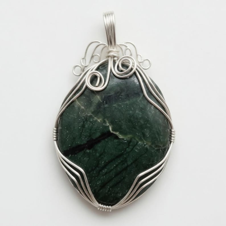 Unique certified green jade wire-wrapped crystal necklace photographed to highlight the 90-100 carat gemstone base and custom wire structure resting on a light surface. The natural green stone features individual variations that make each piece distinct. Designed as handmade artisan jewelry, this wrapped necklace works well as a statement accessory for spiritual fashion, casual styling, special events, and thoughtful gifts for crystal lovers looking for something unique.