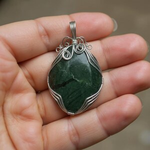 Close-up photograph of a certified green jade wire-wrapped crystal necklace showing the polished 90-100 carat gemstone base mounted with detailed silver wirework across the stone. The pendant rests securely on a neutral textured background. This natural crystal pendant makes an elegant statement accessory for bohemian fashion, everyday wear, crystal collections, and meaningful gifts for family and friends seeking unique artisan jewelry pieces with natural earthy character.