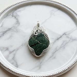 Artisan-crafted green jade wire necklace featuring a polished 90-100 carat crystal secured in a detailed silver wire framework with elegant loop accents. The unique design creates a striking appearance across the dark green stone surface, adding texture and elegance to the handcrafted pendant. This certified natural healing jewelry piece is suitable for everyday wear, spiritual outfits, statement jewelry styling, special occasions, and gift giving for someone special.