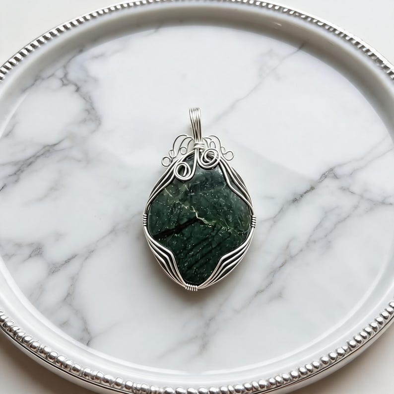 Artisan-crafted green jade wire necklace featuring a polished 90-100 carat crystal secured in a detailed silver wire framework with elegant loop accents. The unique design creates a striking appearance across the dark green stone surface, adding texture and elegance to the handcrafted pendant. This certified natural healing jewelry piece is suitable for everyday wear, spiritual outfits, statement jewelry styling, special occasions, and gift giving for someone special.