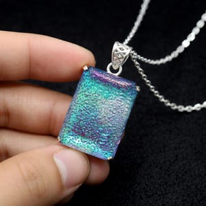 May include: A rectangular pendant featuring a textured, iridescent surface with shifting hues of teal, blue, and purple. The pendant is encased in a silver-toned metal setting with a decorative bail and hangs from a matching silver-toned link chain.