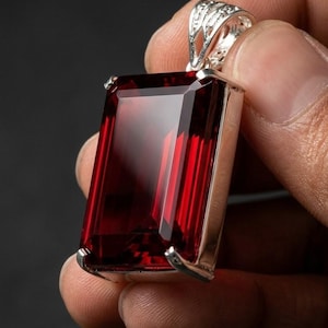 May include: A rectangular, deep red gemstone pendant set in a polished silver-toned metal frame. The stone features a faceted emerald cut, and the pendant includes an ornate, filigree-style silver bail at the top for attaching to a chain.
