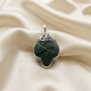 Unique certified green jade crystal wire necklace featuring a natural 90-100 carat gemstone base with precise wire-wrapped strands and decorative top swirls. The polished stone is framed by an artistic silver-tone setting, creating a refined and symbolic handcrafted appearance. This gemstone pendant complements spiritual, bohemian, casual, or elegant fashion and makes a distinctive addition to any collection. Natural variations give every piece unique character and beauty.