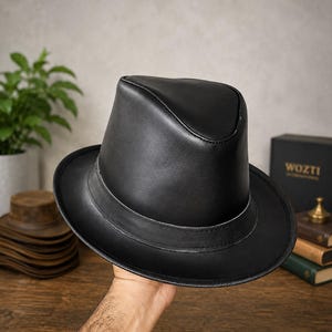 May include: A black leather fedora hat with a structured crown and a wide brim. The hat features a smooth, matte finish and a matching leather band encircling the base of the crown, with visible stitching along the edges of the brim and band.