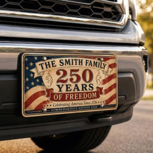 May include: A rectangular metal license plate featuring a vintage American flag design. The text reads THE SMITH FAMILY 250 YEARS OF FREEDOM, Celebrating America Since 1776, and COMMEMORATIVE EDITION 2026. The plate is mounted on the front bumper of a vehicle.