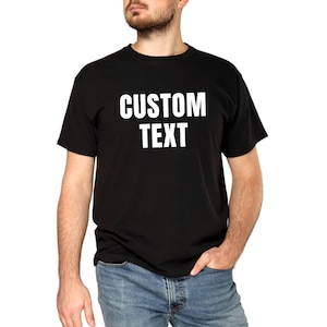 May include: A black short-sleeved t-shirt featuring the words CUSTOM TEXT printed in bold white capital letters across the chest. The garment is paired with blue denim jeans, showcasing a standard crew neck design and a classic fit silhouette.