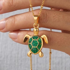 Op de afbeelding: Een goudkleurige ketting met een schildpadhanger met een textuurrijke, glinsterende groene inleg op het schild. De hanger hangt aan een fijne goudkleurige ketting met kleine, gelijkmatig verdeelde bolletjes in het ontwerp.