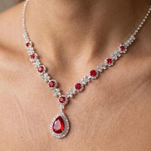 Può includere: Una collana color argento con un ciondolo centrale a forma di pera con pietra rossa circondata da piccoli cristalli trasparenti. La catena è decorata con pietre rosse rotonde alternate e grappoli di cristalli trasparenti lungo tutta la lunghezza.