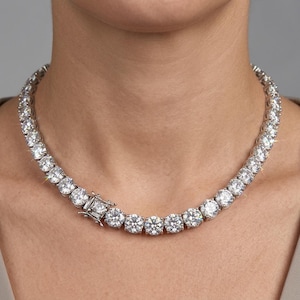 Puede incluir: Un collar de tenis en tono plateado que presenta una hilera continua de piedras redondas, transparentes y facetadas engastadas en un marco metálico. La pieza incluye un cierre de caja seguro en la parte frontal con acabado pulido.