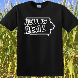 May include: Black t-shirt with the phrase "HELL IS REAL" in white, outlined in black, along with the outline of the state of Iowa. The shirt is set against a background of a cornfield.