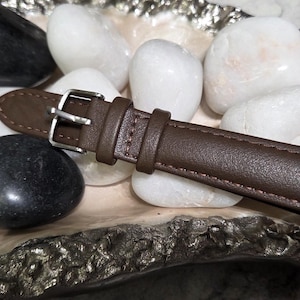 May include: A brown watch strap featuring a silver-toned metal buckle and two matching leather loops. The strap has visible stitching along the edges and is positioned horizontally, showcasing a smooth texture and a classic, functional design.