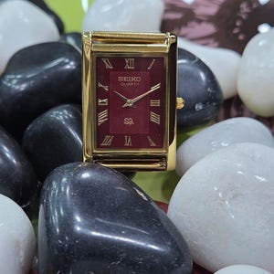 May include: A rectangular gold-tone Seiko Quartz wristwatch featuring a deep red dial with a subtle concentric wave pattern. The watch face displays gold-colored Roman numeral hour markers, gold hands, and the Seiko Quartz logo in gold text.