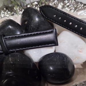 May include: A black watch strap featuring smooth leather-like material with tonal stitching, a silver-toned metal buckle, and two fixed loops. The strap is displayed against a collection of polished black and white smooth stones.