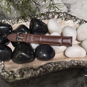May include: A brown leather watch strap with a silver-toned metal buckle resting on a collection of smooth black and white river stones inside an irregular, textured silver-rimmed decorative tray with a light-coloured interior surface.