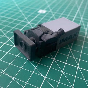 May include: A 3D printed miniature film camera model in black and grey plastic. The device features a rectangular lens hood, a side-mounted viewfinder, and the word IMAX embossed in capital letters on the side of the grey rectangular body.