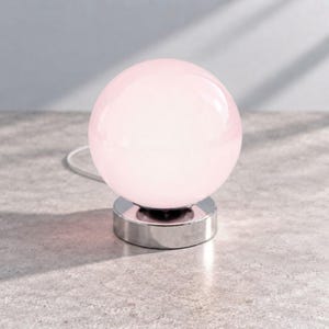 May include: A spherical table lamp featuring a smooth, pale pink glass globe shade. The lamp is supported by a circular, polished silver-tone metal base. A thin white power cord extends from the back of the base, connecting to the electrical source.