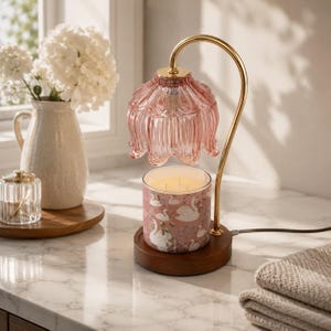 May include: A candle warmer lamp featuring a curved gold metal arm, a pink fluted glass shade, and a dark wood base. A cylindrical candle with a pink label depicting white swans sits beneath the lamp, which emits a warm glow from the bulb.
