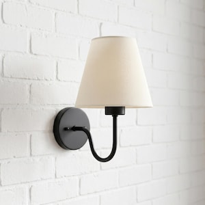 Peut inclure: Une applique murale composée d'une base circulaire noire mate et d'un bras incurvé supportant un abat-jour en tissu blanc cassé de forme conique. Le luminaire est fixé sur un mur en briques blanches, offrant un design épuré et fonctionnel.