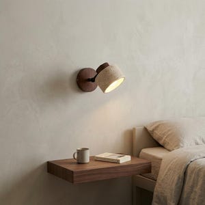 Peut inclure: Une applique murale composée d'une base circulaire en bois foncé et d'un abat-jour cylindrique en pierre beige poreuse. La lampe est fixée au-dessus d'une étagère flottante en bois foncé supportant une tasse en céramique et un livre.