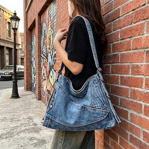 Op de afbeelding: Een mediumblauwe denim schoudertas met een verweerde acid-wash textuur, meerdere buitenzakken met zichtbare naden en zilverkleurige metalen knopen langs het zijpaneel, gedragen aan een brede, geïntegreerde denim schouderband.