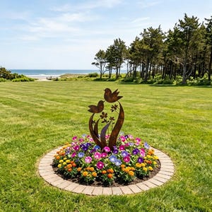 May include: A rust-coloured metal garden sculpture featuring two stylised birds perched atop curved stems and floral shapes, positioned in the centre of a circular flower bed filled with vibrant pink, purple, and orange blossoms.