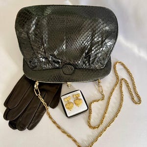 Può includere: Una pochette nera con texture in pelle di serpente e tracolla a catena dorata, un paio di guanti in pelle nera e un piccolo portagioie nero contenente due orecchini dorati a forma di cuore con fiocchi sulla parte superiore.