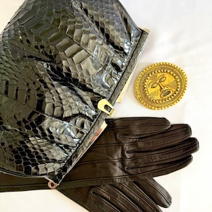 Può includere: Una pochette nera con texture effetto pelle di serpente, telaio e chiusura in metallo dorato, un paio di guanti in pelle marrone scuro con cuciture a vista e una spilla rotonda dorata con un volto in rilievo e bordo testurizzato.