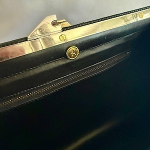 Può includere: L'interno di una borsa nera con una cornice in metallo argentato e un meccanismo di chiusura. Un emblema circolare dorato con una piccola figura è centrato sulla fodera nera, che include anche una tasca orizzontale con cerniera.