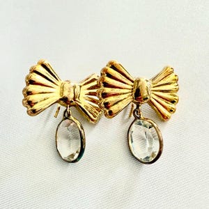 Può includere: Un paio di orecchini in metallo color oro con un design a forma di fiocco e dettagli a coste. Ogni orecchino presenta un ciondolo in cristallo trasparente di forma ovale sospeso al centro del fiocco, fissato con una chiusura a perno.