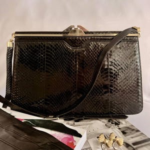 May include: A rectangular black handbag with a glossy snakeskin-like texture, featuring a gold-tone metal frame, a top clasp closure, and a matching shoulder strap. The bag has a structured silhouette with vertical panel detailing on the front.