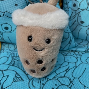 Könnte beinhalten: Ein Plüsch-Boba-Tee-Schlüsselanhänger in Form eines beigen, flauschigen Bechers mit weißem Kunstfellrand, einem kleinen beigen Strohhalm und dunkelbraunen, gestickten Boba-Perlen. Das Gesicht zeigt zwei schwarze Augen und einen lächelnden Mund.