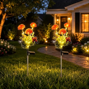 May include: Two solar-powered garden stake lights featuring artificial orange flowers and decorative butterflies. Each stake has a small solar panel attached to the metal pole, with warm yellow LED lights illuminating the flower clusters at night.