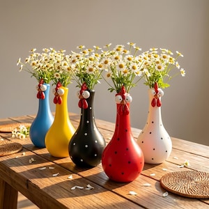 May include: Five ceramic vases shaped like chickens, each holding a bouquet of white daisies. The vases are blue, yellow, black with white dots, red with black dots, and white with black dots. Each features a red comb and white eyes.