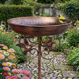 Puede incluir: Un baño para pájaros de metal color cobre con un cuenco ancho y poco profundo sobre un pedestal central. El soporte está decorado con un colibrí y una flor de metal. Un pequeño pájaro amarillo y negro está posado en el borde.