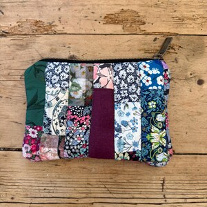 May include: A rectangular zipped pouch constructed from a patchwork of various fabric scraps. The design features a mix of floral, botanical, and solid-coloured textile segments in shades of green, blue, pink, white, and a central deep plum panel.