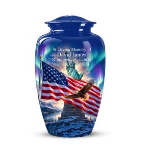 Può includere: Un'urna cineraria blu con una grafica stampata della Statua della Libertà, una bandiera americana e un'aquila calva su uno sfondo di aurore boreali. Il testo recita In Loving Memory of David James 02-08-1998 - 25-11-2025.