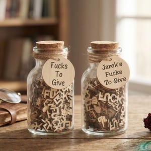 May include: Two clear glass jars with cork stoppers filled with small wooden cutouts of the word "fuck" and middle finger icons. Each jar features a round wooden tag tied with twine; one reads "Fucks To Give" and the other "Jarek's Fucks To Give."