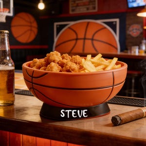 May include: A round orange bowl shaped like a basketball with black lines, featuring a black base with the name STEVE printed in white. The bowl is filled with fried chicken bites and french fries, positioned next to a glass bottle and a cigar.