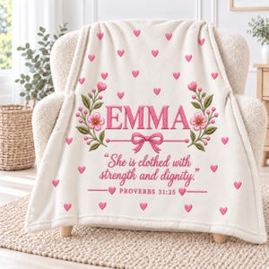 Puede incluir: Una manta de forro polar color crema con el nombre EMMA en letras rosas, flanqueado por ramas florales rosas y un lazo. Debajo, el texto dice "She is clothed with strength and dignity." y "PROVERBS 31:25," rodeado de pequeños corazones rosas.