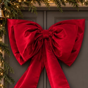 May include: A large, deep red fabric bow with a smooth, slightly lustrous finish. The bow features two wide, voluminous loops and two long, flowing tails that hang vertically. The centre is cinched with a matching red fabric band.