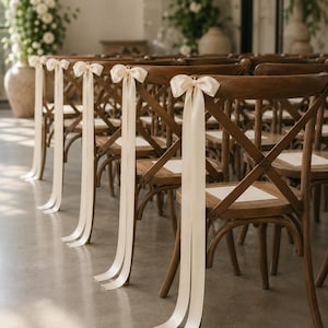 May include: A row of wooden cross-back chairs arranged in a line. Each chair features a smooth, medium-brown wood finish and a light cream-colored satin ribbon tied into a bow at the center of the top rail, with long ribbon tails hanging down.