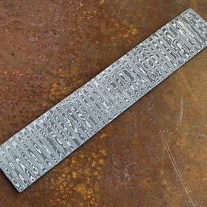 May include: A rectangular metal billet featuring a distinct Damascus steel pattern with alternating dark gray and light silver wavy lines. The surface displays a complex, layered organic design created by the forging of multiple steel alloys.