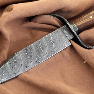 May include: A fixed-blade knife featuring a distinctive Damascus steel blade with a swirling gray and black pattern. The handle is crafted from polished brown wood and secured with two brass pins. A black metal guard separates the blade and handle.