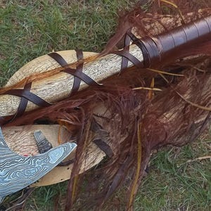 May include: A hand axe featuring a patterned Damascus steel blade with a light-colored wooden handle. The handle is wrapped in dark brown leather strips that crisscross along the shaft and form a solid grip section near the top of the axe head.