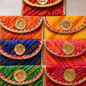 Könnte beinhalten: Eine Sammlung von sieben rechteckigen Stofftaschen mit einem traditionellen Batikmuster in Rot, Orange, Gelb, Blau, Grün und Pink. Jede Tasche hat eine geschwungene Klappe, die mit einem goldenen, kreisförmigen Blumenemblem verziert ist.