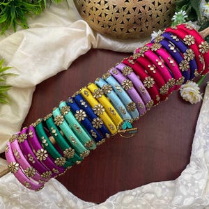 May include: A set of multi-colored silk thread bangles arranged in a row. Each bangle features a smooth wrapped finish in shades of pink, purple, blue, green, and yellow, accented with gold-toned metal flower motifs and small inset mirrors.