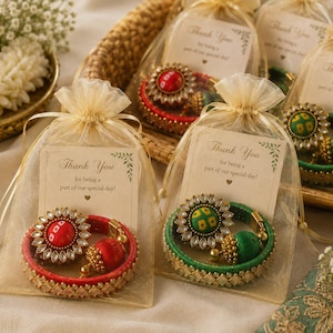 May include: Several sets of round bangles and matching earrings are packaged in individual sheer gold organza drawstring bags. Each bag contains a card with the text Thank You for being a part of our special day. The jewelry features red or green fabric-wrapped bangles with gold-toned accents and floral-shaped embellishments with clear stones.